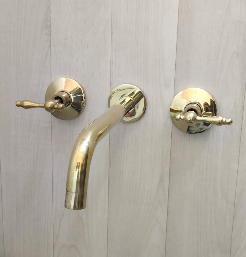 May include: A gold-toned bathroom faucet with two handles and a curved spout. The faucet is mounted on a white wall.