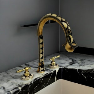Può includere: Un rubinetto con motivo nero e oro con un design decorativo. Il rubinetto ha due maniglie ed è montato su un piano di lavoro in marmo nero.