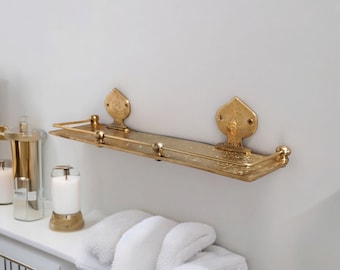 May include: Gold metal shelf with ornate details, mounted on a white wall. The shelf has a decorative design and is perfect for displaying small items.