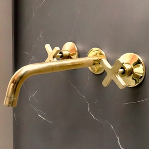 May include: Gold-toned bathroom faucet with two cross-shaped handles mounted on a grey marble wall.