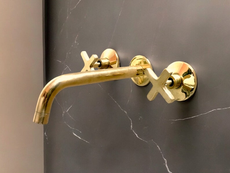 May include: Gold-toned bathroom faucet with two cross-shaped handles mounted on a grey marble wall.