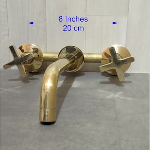 May include: Gold-toned wall-mounted faucet with two cross-shaped handles and a spout. The faucet measures 8 inches or 20 cm wide.