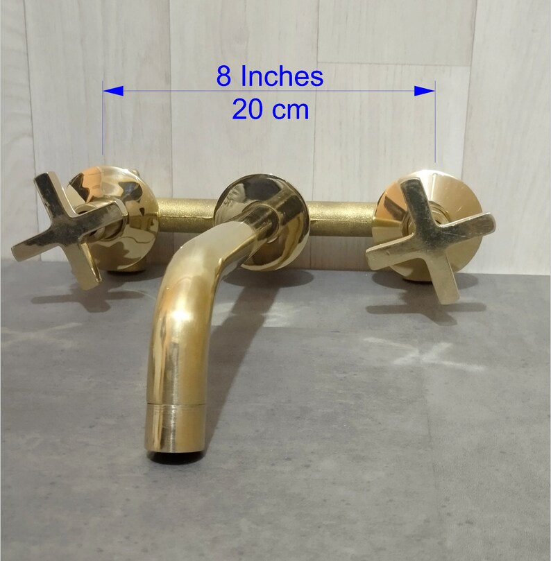 May include: Gold-toned wall-mounted faucet with two cross-shaped handles and a spout. The faucet measures 8 inches or 20 cm wide.