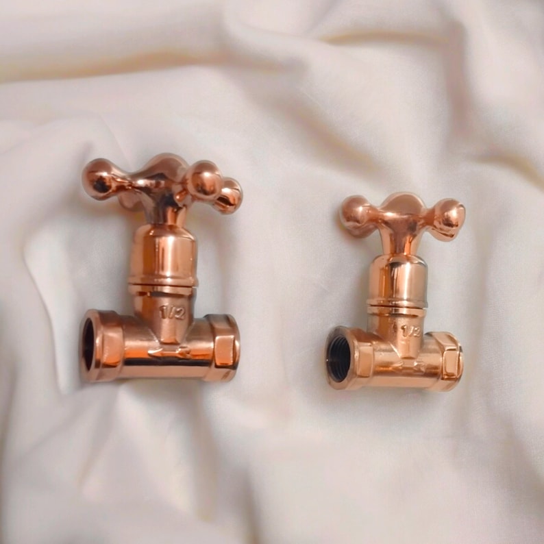 Peut inclure: Deux vannes de plomberie en m&eacute;tal cuivr&eacute; poli avec des poign&eacute;es en forme de croix. Chaque vanne pr&eacute;sente un corps central avec une ouverture filet&eacute;e sur le c&ocirc;t&eacute; et le texte 1/2 grav&eacute; sur le devant, indiquant une taille de raccord de 1/2 pouce.