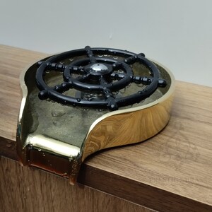 May include: A gold-coloured soap dish featuring a black ship's wheel design. The dish has a curved shape and is on a wooden surface. Water droplets are visible on the dish.