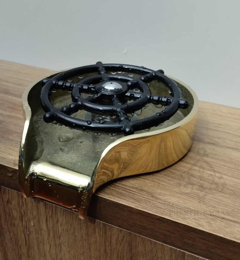 May include: A gold-coloured soap dish featuring a black ship's wheel design. The dish has a curved shape and is on a wooden surface. Water droplets are visible on the dish.