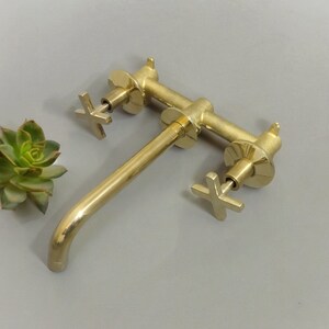 May include: A gold-toned brass faucet with two cross-shaped handles. The faucet has a long spout and is designed for wall mounting.