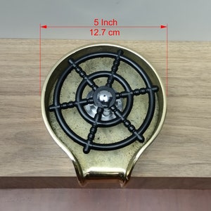May include: A brass ashtray featuring a black ship's wheel design. The ashtray is about 12.7 cm in diameter. It has a rounded shape with a small handle. The ashtray is set on a wooden surface.