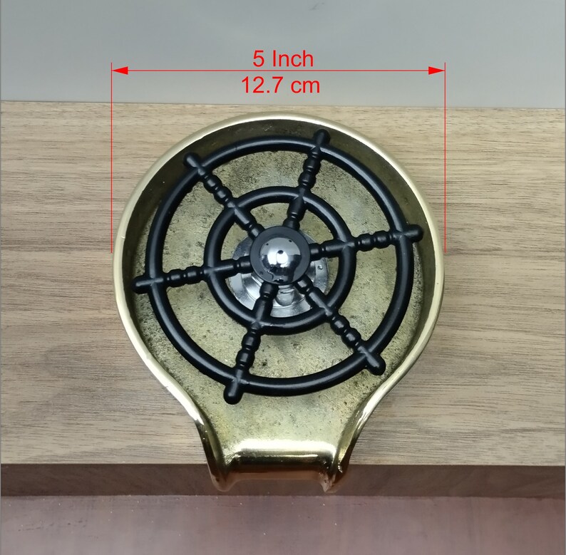 May include: A brass ashtray featuring a black ship's wheel design. The ashtray is about 12.7 cm in diameter. It has a rounded shape with a small handle. The ashtray is set on a wooden surface.