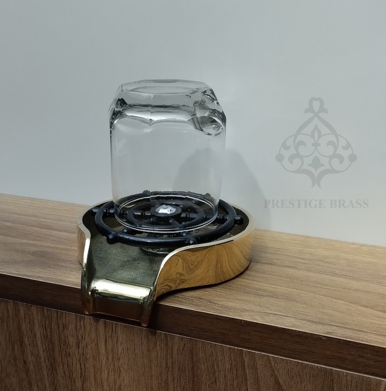 May include: A clear glass tumbler sits upside down on a brass and black bar glass rinser. The rinser has a gold finish and a circular design with a black centre. The product is on a wooden surface.