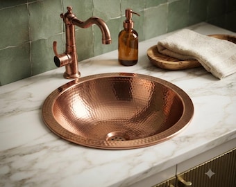 Op de afbeelding: Een ronde, gehamerde koperen wastafel met een metallic afwerking, gecombineerd met een bijpassende koperen kraan. De wastafel is ingebouwd in een wit marmeren aanrechtblad naast een bruine glazen zeepdispenser en een houten dienblad.