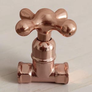 Op de afbeelding: Een gepolijste koperkleurige metalen kraangreep met een klassiek kruisvormig ontwerp met vier afgeronde uiteinden. Het object heeft een glad, reflecterend oppervlak en een cilindrische basis die verbonden is met een horizontaal buisstuk.