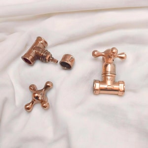 Può includere: Accessori idraulici color rame, tra cui un rubinetto, una valvola e una maniglia, sono disposti su una superficie bianca. Il rubinetto ha una maniglia a forma di croce e la valvola è contrassegnata con "1/2". Gli accessori hanno un'estetica vintage.