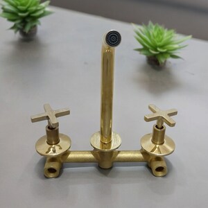 May include: A gold-toned faucet with two cross-shaped handles and a single spout. The faucet is mounted on a white surface.