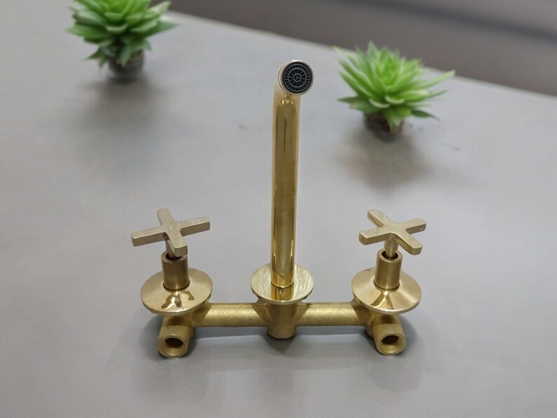 May include: A gold-toned faucet with two cross-shaped handles and a single spout. The faucet is mounted on a white surface.