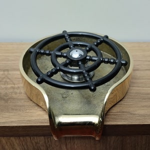 May include: A brass-coloured ashtray featuring a black ship's wheel design. The ashtray has a rounded shape with a small handle. The ship's wheel is centered within the ashtray.