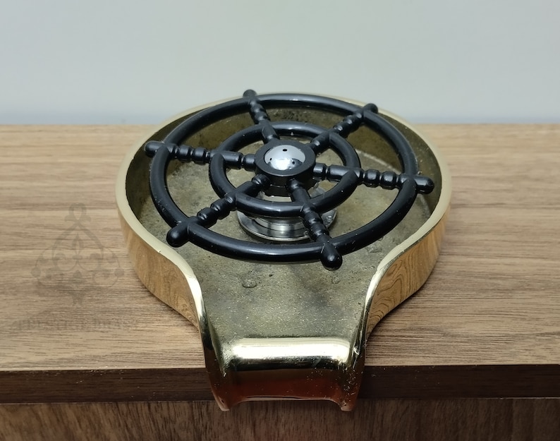 May include: A brass-coloured ashtray featuring a black ship's wheel design. The ashtray has a rounded shape with a small handle. The ship's wheel is centered within the ashtray.