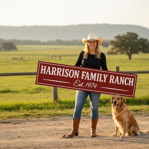 Smiling woman in a cowboy hat holding a custom barn red ranch sign that reads ‘Harrison Family Ranch Est. 1974,’ with a golden retriever sitting beside her on a dirt road overlooking a green pasture, handcrafted by Farmhouse Timber Company.”