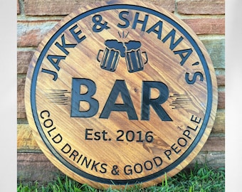 May include: A round wooden sign with a dark brown stained finish and black carved lettering. The text reads JAKE & SHANA'S BAR, Est. 2016, and COLD DRINKS & GOOD PEOPLE. A graphic of two clinking beer mugs is centered above the word BAR.
