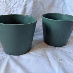 May include: Two green ceramic plant pots. The pots are cylindrical with a slightly flared top. The pots are a muted green color and sit on a white, textured surface. The pots are empty and ready for planting.