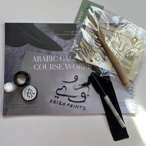 Puede incluir: Un libro de ejercicios de caligrafía árabe titulado Arabic Calligraphy Course Workbook by Faizo Paints, que incluye un juego de plumillas metálicas en una bolsa de plástico, un portaplumas de madera, un tintero negro y un marcador negro.