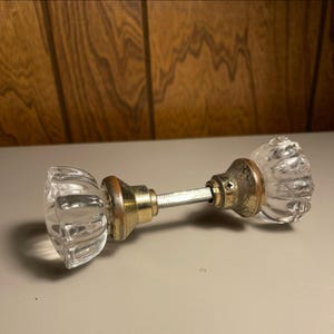 May include: A vintage-style door knob set featuring two clear, fluted glass knobs connected by a central metal spindle. Each knob is attached to a tarnished brass-colored circular base plate, with one base plate showing a small visible screw.
