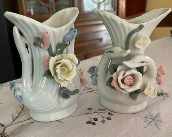 May include: Two white porcelain vases with floral designs. The vases are shaped like swans with their necks curved up. The flowers are in shades of pink, yellow, and blue.