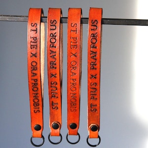 Può includere: Quattro portachiavi in ​​cuoio arancione con anelli metallici. Ogni portachiavi ha inciso il testo "ST PIUS X PRAY FOR US" o "ST PIE X ORA PRO NOBIS".