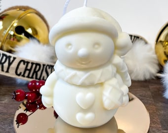Pode incluir: Vela de boneco de neve branco com um ramo de bagas vermelhas e um sino dourado no fundo. O boneco de neve tem uma echarpe vermelha, um chapéu preto e dois corações no peito.