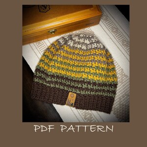 Op de afbeelding: Een gebreide beanie met bruine, groene en gele strepen. De beanie heeft een bruin leren patch met een bergontwerp aan de zijkant. De beanie ligt op een wit en bruin gepatroneerd oppervlak.