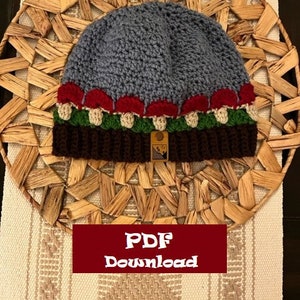 Op de afbeelding: Een gebreide beanie met een paddenstoelontwerp. De muts is blauw met rode en witte paddenstoelen en groene bladeren. De muts heeft een bruine rand en een klein label met een berglogo.