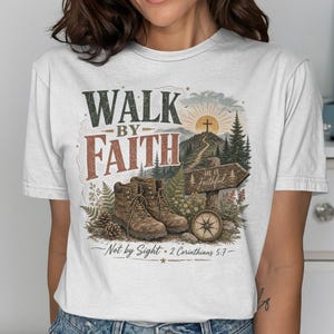 May include: A white short-sleeve t-shirt featuring a graphic print of brown hiking boots, a compass, a wooden sign reading He is Faithful, and a mountain landscape. The text reads WALK BY FAITH and Not by Sight 2 Corinthians 5:7.