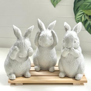May include: Three white ceramic bunny figurines in a 'see no evil, hear no evil, speak no evil' pose, arranged on a small wooden pallet. The bunnies have textured surfaces and are set against a white background with a hint of greenery.