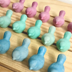 Puede incluir: Una fila de pequeñas figuritas de pájaros de cerámica en varios tonos de rosa, azul y verde. Los pájaros están dispuestos en línea sobre una superficie de madera.