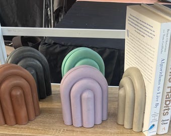 Puede incluir: Un conjunto de sujetalibros decorativos en varios colores, incluyendo marrón, negro, verde y lavanda. Los sujetalibros tienen un diseño arqueado y sostienen dos libros titulados "Atomic Habits" y "Algorithms to Live By".
