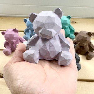 Puede incluir: Una colección de pequeñas figuritas de ositos de peluche geométricos en varios colores pastel. Los osos están hechos de un material liso y texturizado y tienen un diseño simple y moderno.