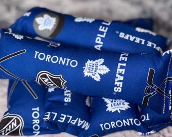 May include: A set of blue fabric bean bags featuring a repeating pattern of the Toronto Maple Leafs logo, the text "TORONTO MAPLE LEAFS", the NHL shield logo, and black hockey stick graphics. The fabric is printed in white and black ink.