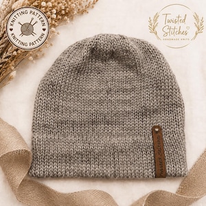 May include: A grey knit beanie featuring a stockinette stitch pattern and a brown leather tag attached to the lower edge. The tag is embossed with the text Twisted Stitches. The beanie has a rounded crown and a simple, un-cuffed bottom edge.
