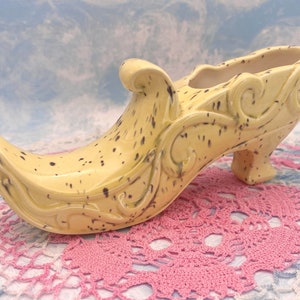 May include: A yellow ceramic shoe planter with a black and white speckled glaze and intricate floral designs. The shoe has a high heel and a pointed toe.