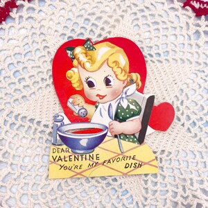 Puede incluir: Tarjeta vintage de San Valentín con una niña de dibujos animados en un marco rojo en forma de corazón. La niña sostiene una cuchara y un salero sobre un tazón de sopa roja. El texto de la tarjeta dice "Querido Valentín Eres mi plato favorito".