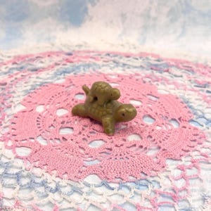 May include: A small, olive-green figurine of a mother turtle with a baby turtle on its back. The turtles are detailed with textured shells. The figurine is set on a pink and white crocheted doily, with a soft blue and white background.