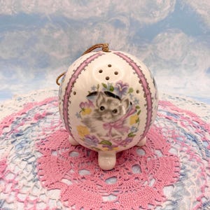 Puede incluir: Adorno esférico de cerámica blanca con una ilustración de gato, detalles de cinta rosa y pequeños agujeros. El adorno tiene tres pequeños pies y un cordón dorado para colgar. Detalles florales en amarillo, azul y morado rodean al gato.