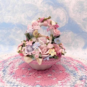 May include: A decorative basket filled with pastel fabric flowers, including roses and other blooms. The basket has a handle decorated with pink ribbons and sits on a pink lace doily, creating a charming, feminine aesthetic.