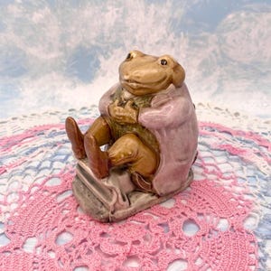 Könnte beinhalten: Keramikfigur einer Froschfigur in einem rosa Mantel und braunen Hosen, sitzend auf einem steinartigen Sockel. Der Frosch hat einen beigefarbenen Körper und hält die Hände zusammen. Ein skurriles Dekorationsstück.