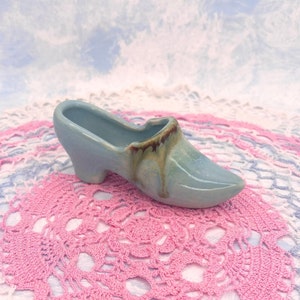 May include: A light blue ceramic shoe shaped planter with brown glaze accents. The planter is sitting on a pink crocheted doily.