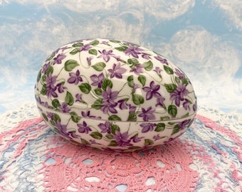 Könnte beinhalten: Eine weiße, eiförmige Schmuckschatulle aus Keramik mit einem floralen Muster aus violetten Blüten und grünen Blättern. Die Schachtel steht auf einer rosa-weißen Häkeldeckchen.