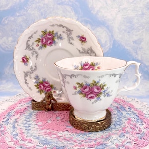 Puede incluir: Un juego de taza y platillo blancos vintage, adornado con rosas rosas y detalles grises. La taza y el platillo descansan sobre soportes de latón ornamentados. El juego se presenta sobre un tapete de encaje rosa y azul, evocando una sensación clásica y elegante.