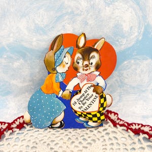 Könnte beinhalten: Vintage Valentinstagskarte mit zwei anthropomorphen Hasen. Einer trägt ein blaues Kleid mit weißen Punkten und eine orangefarbene Jacke. Der andere Hase hält ein herzförmiges Schild mit der Aufschrift "I'd Jump At The Chance To Be Your VALENTINE."