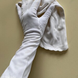 May include: A pair of white elbow-length gloves made of a smooth, opaque fabric. The gloves feature a scalloped edge detail along the wrist opening and a decorative stitched seam running along the back of the hand and fingers for a tailored fit.