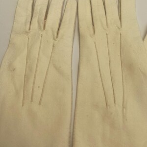 May include: A pair of off-white leather gloves. The gloves have a classic design with visible stitching along the seams and fingers. The gloves are made of a soft, supple material.